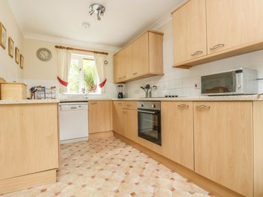 A kitchen with wooden cabinets a window with curtains a dishwasher an oven and a microwave at Cuckoo's Cottage in Falmouth