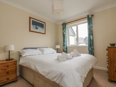 A bedroom with a double bed white bedding rolled towels bedside tables lamps a window with blue floral curtains and a framed picture on the wall at Cuckoos Cottage in Falmouth