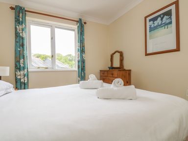 A bedroom with a large bed with white linens and rolled towels near a window with blue floral curtains and a wooden dresser with a mirror at Cuckoo's Cottage in Falmouth