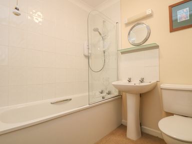 A bathroom with a bathtub shower, pedestal sink, toilet, mirror, and framed picture at Cuckoo's Cottage in Falmouth