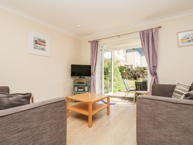 A living room with brown sofas a wooden coffee table and a TV on a metal stand next to open doors leading to a garden at Trelease in Falmouth