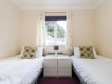 A bedroom with two single beds separated by a small white nightstand with a lamp and a window with white curtains at Trelease in Falmouth