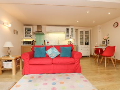 A living room with a red sofa and blue cushions facing a kitchen with white cabinets and a dining area with red chairs at Plum Cottage in Colebrooke near Crediton