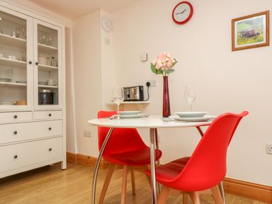 A dining area with a round table set for two with red chairs a white cabinet and a wall clock at Plum Cottage in Colebrooke near Crediton