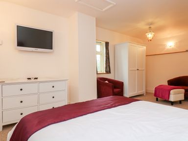 A bedroom with a bed covered in white and red bedding a white dresser with a TV mounted above red chairs a white wardrobe and a footstool with a red cover at Plum Cottage in Colebrooke near Crediton