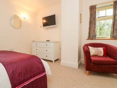 A bedroom with a bed covered in a maroon blanket a white dresser a wall-mounted TV and a maroon armchair with a cushion at Plum Cottage in Colebrooke near Crediton