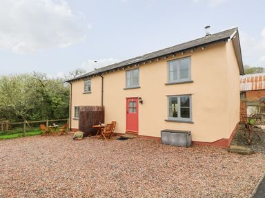 A house with a red door and two sets of wooden tables and chairs on a gravel driveway at Apple Cottage in Colebrooke near Crediton