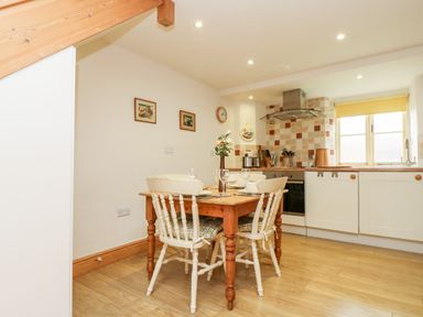 A kitchen with a wooden dining table and four chairs plates and glasses on the table white cabinets and tiled backsplash with a window in the kitchen at Apple Cottage in Colebrooke near Crediton