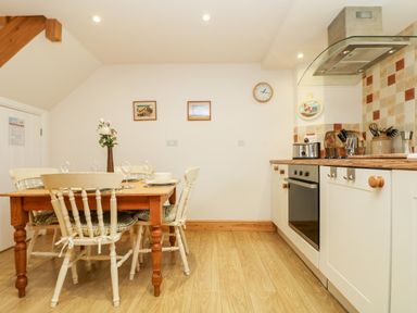 A kitchen and dining area with a wooden table set with plates and glasses and white chairs with cushions at Apple Cottage in Colebrooke near Crediton