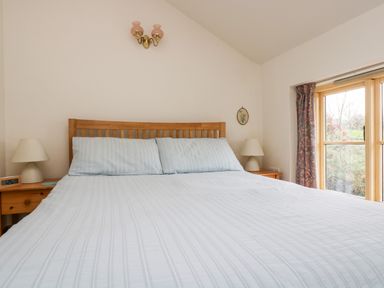 A bedroom with a double bed with light striped bedding two bedside tables with lamps and a window with patterned curtains at Apple Cottage in Colebrooke near Crediton