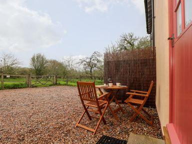 A small outdoor patio with a wooden table and two chairs on a gravel surface next to a red door and fenced garden at Apple Cottage in Colebrooke near Crediton