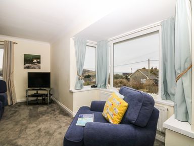 A living room with a blue armchair facing windows with light blue curtains and a television on a black stand at Bethany Bay View in Holywell Bay