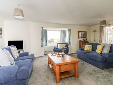 A living room with blue sofas and armchairs around a wooden coffee table with wine glasses and a bottle at Bethany Bay View in Holywell Bay