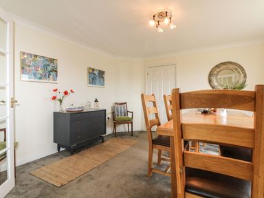 A dining room with wooden chairs and table a black dresser with flowers and artwork on walls at Bethany Bay View in Holywell Bay