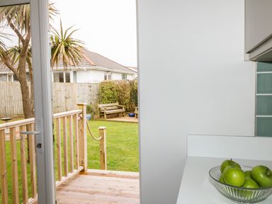 A kitchen counter with a bowl of green apples and a view of a backyard garden with a wooden deck and bench at Bethany Bay View in Holywell Bay