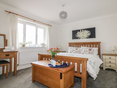 A bedroom with a wooden bed frame a chest at the foot of the bed with flowers on it and a dressing table by a window at Bethany Bay View in Holywell Bay