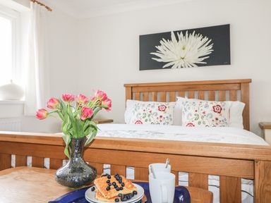 A bedroom with a wooden bed frame floral pillows a tray of waffles with blueberries two mugs and a vase of pink flowers on a table at Bethany Bay View in Holywell Bay