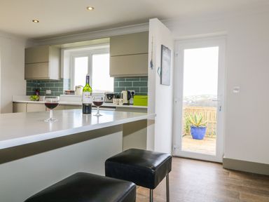 A kitchen with a countertop holding a wine bottle and two glasses two black stools and a door leading outside at Bethany Bay View in Holywell Bay