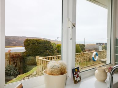 A window with a potted cactus, a ceramic pitcher, and a small decorative tile on the windowsill overlooking a fenced garden with bushes and hills at Bethany Bay View in Holywell Bay