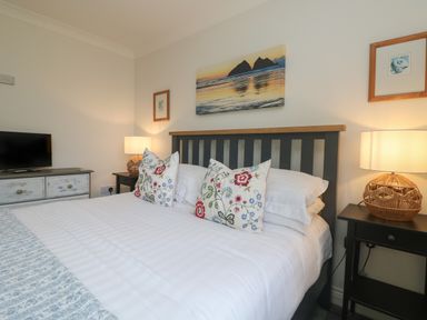 A bedroom with a bed with floral pillows two bedside tables with lamps a chest of drawers and a wall painting at Bethany Bay View in Holywell Bay