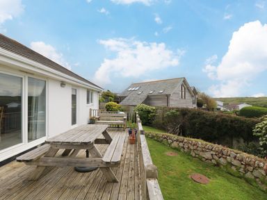 A wooden deck with picnic table and bench overlooking a garden with stone wall and neighboring houses at Bethany Bay View in Holywell Bay