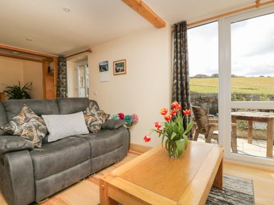 A living room with a grey sofa with patterned cushions a wooden coffee table with red tulips and a view of a patio and fields at Mynheer Farm Barn Nr Portreath