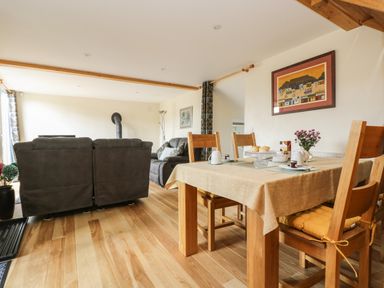 A dining area with a wooden table and chairs next to a living room with two grey sofas and a stove at Mynheer Farm Barn Nr Portreath