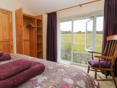 A bedroom with a bed, wooden wardrobe, wooden chair, sliding glass door with purple curtains, and a view of a grassy field at Mynheer Farm Barn Nr Portreath
