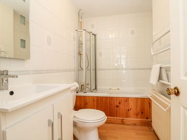 A bathroom with a white sink, toilet, bathtub with a glass shower screen, wooden paneling, and white tiled walls at Seabien in Falmouth