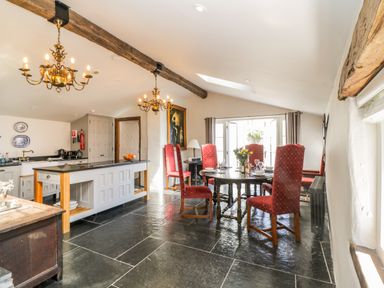 A dining area with a round table and high-backed chairs and a kitchen with an island under wooden ceiling beams at Glanville House in Mevagissey