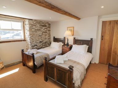 A bedroom with two wooden single beds with white bedding and plaid throws a wooden nightstand with a lamp between the beds a stone wall a window with blinds a wooden door and a carpeted floor at Glanville House in Mevagissey