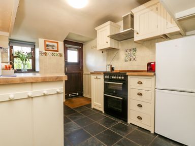 A kitchen with cream cabinets a black oven a white refrigerator a wooden door and a small window with flowers at Gurnard's Cottage in St Ives