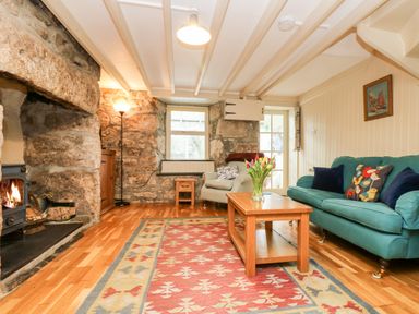 A living room with stone walls a wood burning stove a teal sofa an armchair and a wooden coffee table with flowers at Gurnard's Cottage in St Ives