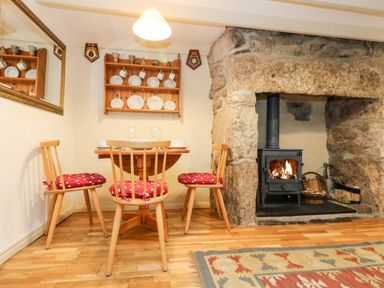 A dining area with a wooden table and three chairs with red cushions near a stone fireplace with a wood-burning stove at Gurnard's Cottage in St Ives