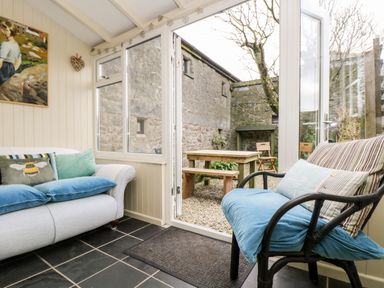 A sitting area with a sofa and chair next to a glass door leading to an outdoor space with a wooden table and benches at Gurnards Cottage in St Ives