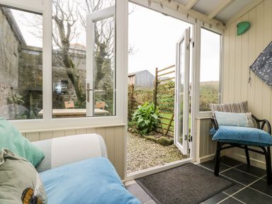 A sunroom with a sofa and a chair facing a glass door opening to a garden with gravel and plants at Gurnards Cottage in St Ives