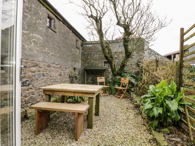An outdoor patio with wooden table and benches gravel ground stone walls and plants at Gurnards Cottage in St Ives
