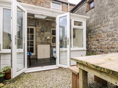 An outdoor patio area with a wooden picnic table and open white-framed glass doors leading into a room with stone walls and tiled floor at Gurnard's Cottage in St Ives
