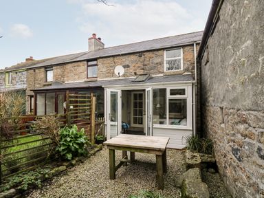 A garden with a wooden table and stone walls outside a stone house with white framed glass doors at Gurnard's Cottage in St Ives