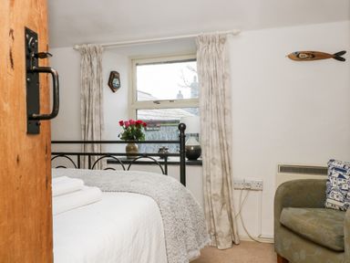 A bedroom with a bed, folded towels, a window with curtains, a vase with flowers, a lamp, an armchair, and wall decorations at Gurnard's Cottage in St Ives