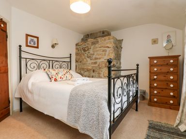A bedroom with a black metal bed frame white bedding stone wall wooden chest of drawers and framed pictures at Gurnards Cottage in St Ives