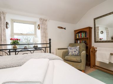 A bedroom with a bed, an armchair, a bookshelf, a mirror, and a window with curtains at Gurnard's Cottage in St Ives