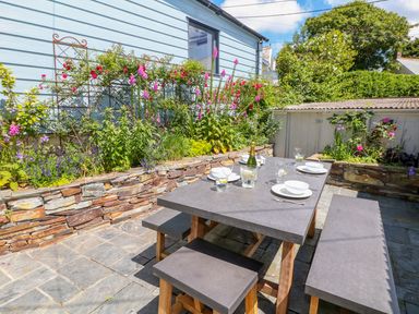An outdoor dining area with a table set with plates and glasses next to a flower bed and light blue building at Parwin in Tintagel