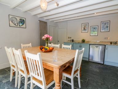 A dining room with a large wooden table surrounded by white chairs a bowl of fruit and flowers on the table blue cabinets with a dishwasher and framed pictures on the wall at Parwin in Tintagel