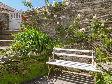 A white bench with roses climbing a stone wall next to stone steps and green plants at Parwin in Tintagel