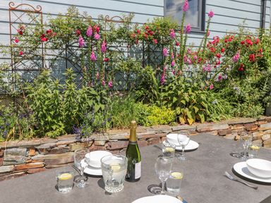 An outdoor dining table set with plates glasses and a bottle in front of a flower bed with pink and red flowers at Parwin in Tintagel