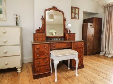 A wooden dressing table with a mirror and a cushioned stool beside a white chest of drawers and a wooden wardrobe at Parwin in Tintagel