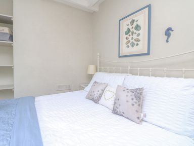 A bedroom with a bed with white and blue linens three cushions a bedside table with a lamp and wall art at Parwin in Tintagel
