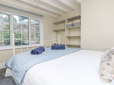 A bedroom with a white metal bed frame a folded blue blanket blue towels on the bed shelves with storage baskets and a window showing an outside stone wall at Parwin in Tintagel