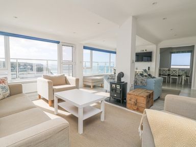 A sitting area with beige and blue armchairs a white coffee table a small stove and large windows overlooking the sea at St Ives View in Porthtowan
